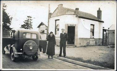 This image shows a very well dressed elderly woman wearing a fur collared long coat and hat, beside a motor car, which is parked on the road.  A man, dressed in a three-piece suit is standing on the edge of the footpath behind her.  The gutter is paved. The back of the car is visible with the number plate 193733.  Both doors are wide open and a large spare wheel is attached to the back of the car by a frame.  A small building with a tiled roof, and bricks part way up the walls is behind the couple.  The front door is open and POST OFFICE is printed above the door way. Two windows can be seen either side of the door.  Both have printing at the bottom of the window which reads “Post & Telegraph Office”.  1937 is printed in large numbers on the bricks on the right-hand side of the building.  Part of a telephone box can be seen behind the post office and part of another building can be seen beside it.  Part of a building with a fence in front can be seen on the left-hand side of the post office.  Two large trees can be seen in front of the motor car.  