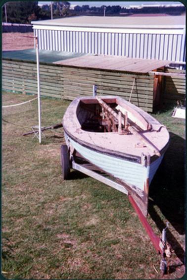 This image shows a small fishing boat on a trailer, parked on grass.  There is a timber structure with a flat iron roof behind the boat and a large shed behind that.  Buildings and trees can be seen in the far background.  There is a thin white pole to the right of the boat.  