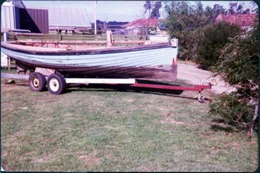 This image shows a wooden fishing boat, side-on, on a four-wheeled trailer, parked on grass.  Some netting can be seen over the side of the boat.  There are sheds in the background and red-roofed buildings on the right-hand side of the photograph.  A line of shrubs is on the right-hand side of the photograph between the boat and the building.   