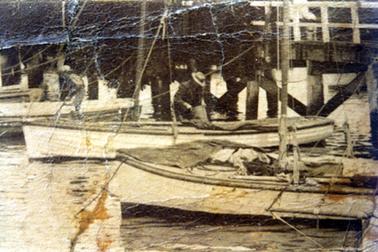 This image shows three fishing boats tied up below the pier.  The boat at the far left and the middle boat have a man each standing at their side.  Both men are wearing hats.  Tall masts can be seen on the front and middle boats.  The front boat is filled with nets and fishing gear.  