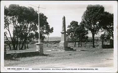 This image shows the soldiers’ memorial at Hastings, a tall narrow stone statue with a pointed top. A low brick wall is on either side, and on the left, set into the brickwork, is a mast. In the background are Western Port Bay and French Island.