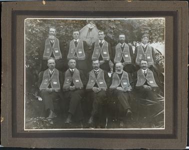 This image shows a group of ten men sitting in two rows of five, wearing Orange Lodge Regalia. They are all wearing suits and ties and the regalia is around the neck and across the shoulders joined by a large toggle.  They all have the number 219 on the regalia with a different initial above it.  The man in the centre of the front row has an extra fringed collar on his regalia.  The men are all ages from young to elderly.  Three of the man are clean shaven and rest have moustaches and/or beards.  There is a flag with the Union Jack and the word LIBERTY printed on it behind the back row of men.  There are a lot of tree branches and leaves behind them.    