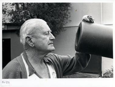 This image shows a side-on photo of a man, from the chest up, looking into the barrel of a gun. He is wearing a Western Port Festival T-shirt and a cardigan.  He has a rag in his left hand which is on the gun barrel .  Part of a wall can be seen in the background.  There is a tree in front of the building.  