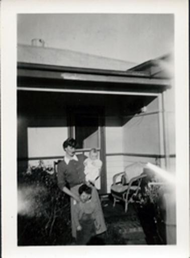 This image shows a woman holding a baby with a small boy standing in front of her posing for a photograph in front of a house.  The woman is wearing a skirt and cardigan with the white collar of her blouse showing.  She has short dark hair.  The baby is dressed in white and the little boy is wearing a long sleeve shirt underneath a pair of overalls.  Part of the house and verandah can be seen behind them.  There is a cane chair with cushions on the right-hand side of the verandah and small shrubs are on either side of the image.  Patches of light have been let into both sides of the image.  
