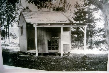 This image shows a small galvanised iron building with a three post awning at the front.  A wooden door is on the left of the building and the sign ‘POST OFFICE’ with mail boxes underneath, is at the centre of the building.  A small white door is on the right-hand side.  A small window is on the side of the building.  Tall trees are beside and behind the building.  