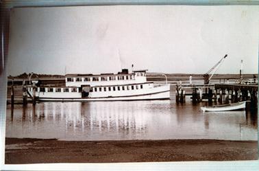 This image shows a double deck boat sitting in the water beside a jetty with the shore in the foreground. The boat has windows right along both decks.  Two people are standing on the jetty, one dressed in white, the other dressed in black.  A small crane is attached to the jetty.  A small boat is tied up to the jetty on the far right-hand side.  Land can be seen in the background.  A dark coloured tank is visible above the boat in the centre of the image.  