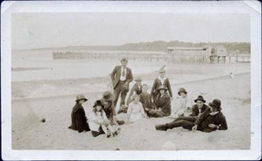 This image shows a group of twelve people on the sand with a jetty, boat sheds and water in the background.  There are six females and six males.  One man is standing, two are kneeling, two are half lying and the remainder are sitting.  All except three are wearing hats.  Two of the woman are wearing coats and all the men are in suits.  Land and houses can be seen on the far background.  