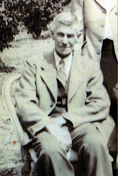 This image shows an elderly man sitting outdoors on a cane chair.  He is wearing a light coloured three piece suit, white shirt and striped tie. He has a slight smile on his face.  Part of a woman can be seen standing behind him to the right. She is wearing a dark coloured skirt, white blouse and light coloured unbuttoned coat.  Fruit trees can be seen to the left of the man.  