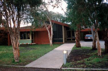 This image shows a building with large glass panels beneath a slanting roof and double glass doors to the right.  A motor vehicle is parked in front of a carport on the far right.  A colourful flower garden is in front of the building on the left-hand side.  A concrete path leads up to the doors.  Grass is on either side of the path and six gum trees are in the foreground.  A short light pole is on the right-hand side of the path in the foreground.  
