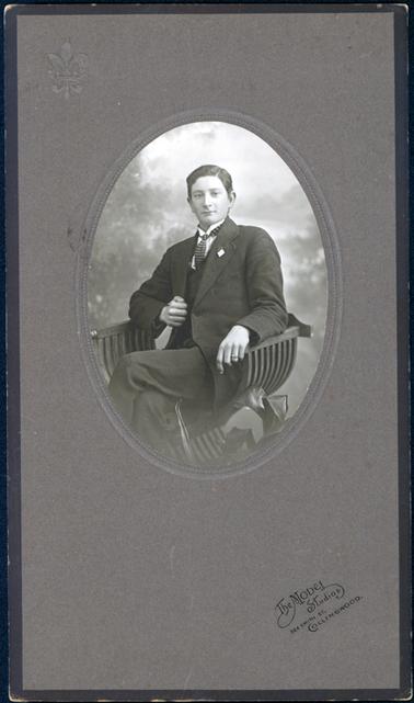 This oval image is a studio portrait of a very well dressed young man sitting in a beautiful curved wooden chair. He is wearing a three piece suit, white shirt and striped tie.  The collar on his shirt is very high with a tiny turndown.  His arms are resting on the arms of the chair. His right hand  is holding the side of his coat.  There is a ring on the fourth finger of his left hand.  His hair is parted on the left side.  Leaves of a pot plant can be seen at the bottom on the right-hand side of the image.  The photographers details are stamped on the mounting board.  