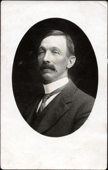 This oval image shows a portrait of a man from the chest up.  The photo is taken slightly side-on.  He is wearing a three-piece suit.  His white shirt has a very high collar.  He has a large moustache and neatly combed hair with a part on the left-hand side.  The photographer’s name is embossed on the bottom right-hand side of the mounting.  