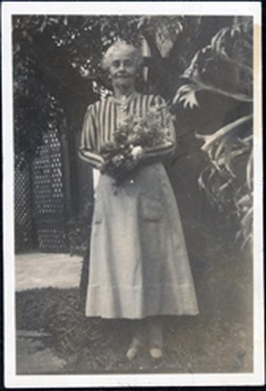 This image shows an elderly, grey haired woman standing posing for a full-length photo.  She is holding a bunch of flowers and standing on grass in front of a large tree.  There is a large flowering plant on the right-hand side of the photograph and a lattice construction on concrete on the left-hand side.  She is wearing a striped long sleeved blouse and a plain coloured skirt with a large hem and two large pockets.