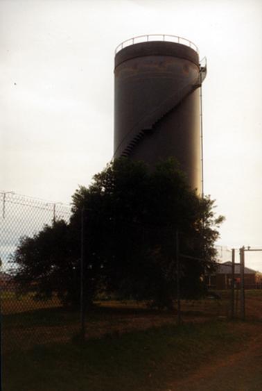 This photograph shows a large water tower at the centre of the image with trees in the front and houses in the background.  The tower has steps going from the bottom left of the tower, across the middle to the top right.  A railing is around the top of the tower. A tall wire netting fence is in the foreground.  