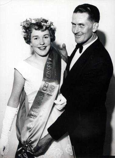 This image shows a young woman being presented with a sash by a man.  She is wearing a round neck, sleeveless ball gown, elbow-length gloves, drop earrings and a wreath of flowers in her short curly hair.  A large brooch/clasp holds the sash together. The man is dressed in a black suit, white shirt and black bow tie.  They are both smiling and looking at the camera.  