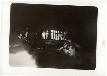 This image shows a cottage engulfed in flames and smoke.  The image is very dark, but the cottage can easily be defined with the flames inside.  A silhouette of trees can be seen on the left-hand side.  