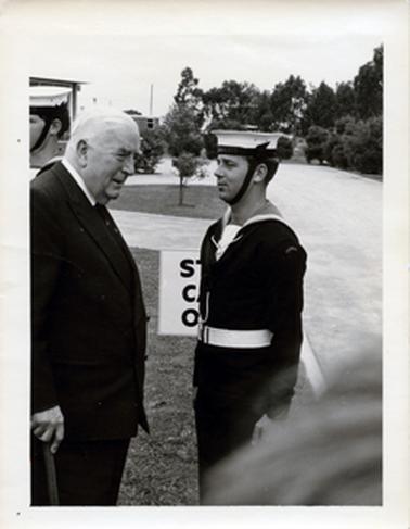 This image shows two men standing, facing each other.  The elderly man on the left is dressed in a dark suit, white shirt and tie.  He has grey hair and is holding a walking stick in his right hand.  The young man on the right is dressed in Navy uniform and standing to attention.  Part of another sailor can be seen on the far left-hand side of the image.  Three black letters, SCO, on a board on the grass is visible behind the sailor. A street, footpath and trees are in the background.   