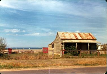 This image shows a small weatherboard  house with a galvanized iron roof.  The roof has faded pale red stripes and some rust.  There is a verandah at the front with four posts.  A door and window can be seen at the front and a small window on the left-hand side of the house.  A large shrub is at the front of the house which sits behind a post and wire netting fence.  A small gate is in the centre of the fence.  Two small red structures can be seen in the back yard and part of another building is on the far right-hand side.  Water is in the background and a road is in the foreground.  