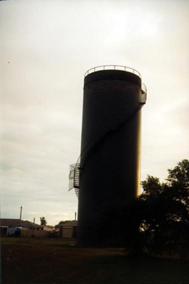 This photograph shows a large water tower at the centre of the image with trees to the right and houses to the left.  The tower has steps going from the bottom left of the tower, across the middle to the top right.  A railing is around the top of the tower. 