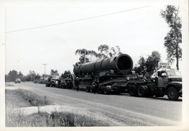 This image shows a very large cylinder on a semitrailer with a truck at the rear.  Another truck is on the road in the distance.  Two men are walking away from each other beside the vehicles.  A large sign is on the top of the semitrailer roof which reads: “DANGER-LONG-WIDE-LOAD”.  Trees can be seen behind the vehicles and grass and a bitumen road is in the foreground.  Two driveways with small posts can be seen across the footpath.  An electricity wife is across the top of the image.   