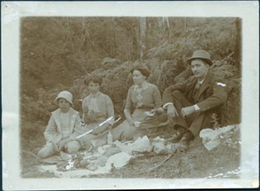 This image shows a family group sitting in bush land enjoying an picnic.  The party consists of a boy on the left, two woman and a man on the right of the photograph.  The boy is wearing a jacket over a white shirt,  long shorts and a hat.  The two ladies are very well dressed with high lace collars and their hair done up.  They each wear a brooch at the base of their necks.  They both have a tea cup in their right hands and the woman on the right has a sandwich in her other hand.  The man is dressed in a three piece suit, tie, white shirt and a hat.  Food is spread out on the ground in front of them.  Bracken fern and tall trees are behind them.