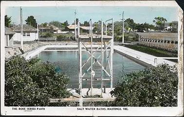The image is a colour postcard of the Salt Water Baths in Hastings. In the centre of the photograph is a high diving board.  There is no one in the pool.  The bathing pavilion is on the right-hand side and houses are on the left and rear of the photograph. Two large shrubs are in the foreground on the left and the right of the diving board.  The printing on the bottom is as follows: “The Rose Series P4378” and “Salt Water Baths, Hastings, Vic.”