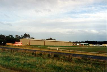 This image shows a very long shed beside green grass and a smaller shed behind it to the right.  A number of small aircraft is sitting on the grass.  Green grass is in the foreground.  Post and wire fences are on either side of a bitumen road and a curved gravel road is running down beside the large building.  Tall trees are in the background.  Red signs with white printing which reads: “Peninsula Aero Club Westernport Airfield” is on the left-hand side beside the bitumen road.  