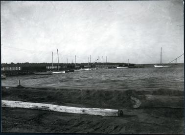 This image is taken from the foreshore looking towards the water.  There is a long jetty commencing on the left-hand side of the image, stretching out into the water.  Several boats are spread out alongside the jetty, all with tall masts.  Two boats, on the right-hand side of the photograph can be seen close to the jetty.  A large pole is lying on the ground in the foreground and a large log is to the right of the pole.  A large line of trees can be seen behind the jetty.  The image is very dull and the sky looks to be heavily overcast.  