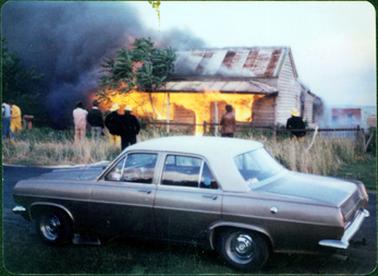 This image shows a burning cottage with the entire front section engulfed in flames. Firemen and other people can be seen standing on the footpath.  A lot of black smoke can be seen billowing to the left-hand side of the image.  A fire hose can be seen across the footpath and over the fence beside a fireman.  A small tree is in front of the verandah.  A post and wire fence is across the front of the cottage.  Long grass is on the footpath and a car is parked on the street in the foreground.  