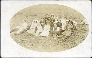 This is an oval photograph of a group of nineteen people.  There are eight men and eleven women.  Everyone is well dressed and all except six are wearing hats.  There is a hat and coat on the ground on the right-hand side and a pile of hats on the grass at the top right-hand side.  The man in the back row,on the right is holding his hat in his right hand.  Two girls are wearing a white veils.  There are pieces of wood and sticks on the rough grass.  