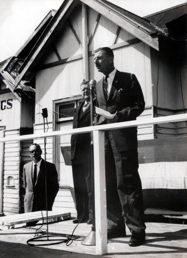 This image shows a man standing on a platform speaking into a microphone, which is on a stand.  He is wearing a suit, white shirt and tie, with a badge in his left lapel.  His right hand is in the pocket of his coat and he is holding a piece of paper in his left hand.  Part of a building can be seen behind him and a white post and railing is in front of him.  A flag is stretched across the rail behind him.  Another man, dressed in a three piece suit, is standing slightly behind him and to his right.  A second microphone, on a stand, is in front of him.  A third man, dressed in a suit and wearing spectacles, is standing on the far left-hand side of the image on ground level.  