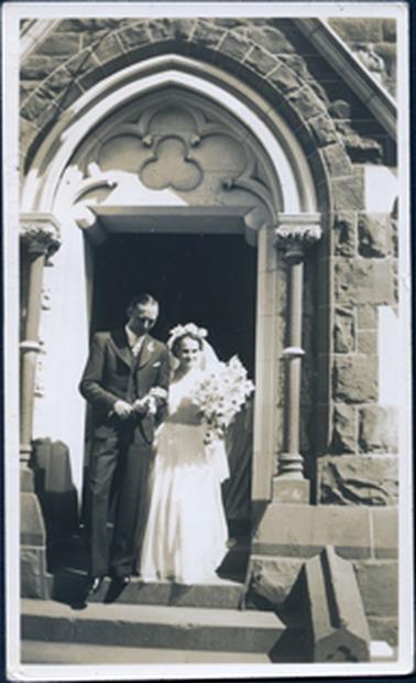 This image shows a bride and groom standing in the doorway of a church at the top of the steps .  The bride has a long, white dress, flowers on the top of her head and a three-quarter length veil.  She is  carrying a large bouquet of flowers.  The groom is wearing a three-piece suit and has a flower pinned to his left lapel.  The bride is holding his left arm.  The church has large sandstone bricks and a carved pattern in the arch.  There are two pillars with ornate tops either side of the steps.  