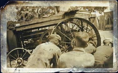 This image shows the top half of four men with their backs to the camera, in front of an old tractor.  Three of the men are wearing caps.  The side view of the tractor shows one small steel wheel at the front and one very large steel wheel at the back.  Three wooden boards are across the top of the frame.  Part of a building with a wooden frame beside it can be seen on the far right-hand side.  Tree branches can be seen in the background. 