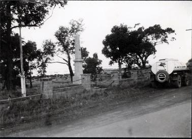 This image shows a monument in park land behind a fence.  The fence has high brick posts with rope or chain strung between them and a lower brick wall underneath.  A flag pole stands on the left-hand side of the image.  There are several trees in the grounds and water can be seen in the background.  A street is in the foreground.  A fuel tanker, with the words PLUME printed on the side is parked on the side of the road beside the fence.  A winged horse and the word VACUUM is printed on the back of the fuel tanker.  