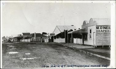 This image is of a street lined with shops.  Some of the shops have verandahs with posts over the footpath.      Writing can be seen on some of the shops.  There are several fences both in front of and between the buildings.  The street is earth with puddles of water in various places.  The inscription on the front of the image reads: “1914 Main St. Hastings (now Marine Pde)”.
