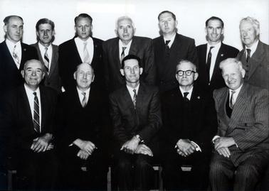 This image shows a group of twelve men, in two rows, posing for a photo.  There are seven men standing in the back row and five men sitting on chairs, with their hands crossed on their laps, at the front.  All men are wearing suits and ties.  
