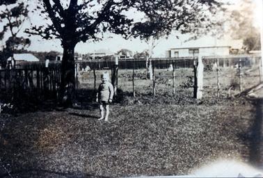 This image shows a small boy standing on lawn smiling for the camera.  He is dressed in shorts and a long sleeved pullover and has bare feet.  A large tree is to his right in the corner of the yard.  Part of a wooden fence can be seen behind the tree.  A post and wire fence is behind him.  An empty paddock with a tall tree in the centre is behind the boy and a long wooden fence stretches across the width of the picture.  Houses can be seen behind the fence.   