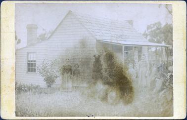 This image shows a small timber cottage with a shingle roof.  There is a verandah with four posts at the front.  A window can be seen on the left-hand side of the house with a large chimney directly above it.  Part of another chimney can be seen on the opposite side of the cottage.  A family of five is standing in front of the cottage looking at the camera.  There are flowers growing on the left-hand side and large shrubs can be seen on the right.  Large trees are in the background, on the right-hand side of the image.  There is long grass in the foreground.  