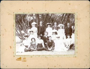 This image shows a group of sixteen men, women and children posing for a photograph in front of trees.  There are six people in the back row, four people kneeling in the middle row and six people sitting on the grass in the front row.  Everyone is beautifully dressed in 19th century clothes.  All the women are wearing hats and the men are in three piece suits.  The group consists of several generations. 