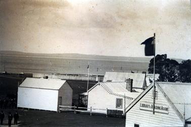 This image shows a group of several weatherboard buildings.  The building in the foreground has the sign  ‘Library & Reading Room’ attached near the roofline and an Australian flag flies from a flagpole attached to the building.  Water is in the background and land can be seen in the far background.  The top of a group of trees is on the far right-hand side of the image.  A group of men can be seen walking at the bottom left-hand side of the image. 