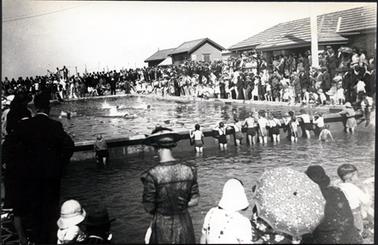 This image shows a large swimming pool surrounded by men, women and children.  There is a row of children standing in another shallow pool with their backs to the camera.  Men women and children are sitting and standing in the foreground with their backs to the camera.  One woman is holding a floral umbrella.  Everyone is watching the swimmers in the pool who are swimming in marked lanes.  Buildings are along the right-hand side.  