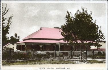This image shows a lovely old home with a red roof and verandah all around.  There is a small building at the rear on the left-hand side of the home.  There is a front fence and part of a side fence can be seen between another building.  A concrete footpath, grass and the street is in the foreground.  A tree is on the footpath surrounded by a boxed support frame.  There are large trees to the left and rear of the image. A flower garden has been planted across the front, just outside of the front fence  Printed at the bottom of the postcard is: “Private Hospital, Somerville. Vic.”.