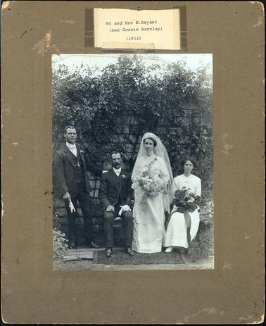 This image shows a wedding group of four people.  The bride is standing and wearing a long dress to the ground with long sleeves and very high lace at the neck. She is wearing a brooch at the base of the neck.  Her hair is swept up underneath a headpiece and veil.  She is holding a very large bouquet.  The groom is seated beside her.  He has a moustache, neatly combed hair and is wearing a three-piece suit with a bow tie.  He is holding his gloves in his left hand.  The best man is wearing similar attire, holding his gloves in his right hand and his left hand is resting on the groom’s shoulder.  The bridesmaid is sitting beside the bride.  She is wearing a long dress with a round neck and a brooch at the front.  She is holding a dark coloured bouquet with dark ribbon.  She has a part in the middle of her hair and is wearing spectacles.  They are posing in front of a weather-board structure which is covered with a creeper.  