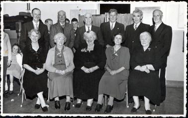 This image shows a group of eleven elderly men and woman posing for a photograph.  There are six men standing in the back row and five women sitting on chairs in front of them.  Everyone is very well dressed.  The men all have three-piece suits, white shirts and ties and the women all have a brooch pinned to their plain coloured dresses.  A little girl can be seen sitting on a chair to the left of the group and four other people can be partly seen standing.  A material screen is behind the men.  