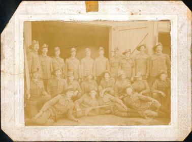 This image shows a group of soldiers in three rows, posing for a photograph.  There are ten men standing at the back, eight men kneeling and crouching in the middle row and four men half-lying on the ground at the front.  They are dressed in full uniform with slouch hats.  Two men are holding small poles with a folded banner/flag.  Part of a shed can be seen behind them with large open wooden doors. The cardboard mounting is embossed. 