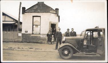 This image shows a group of people standing outside a post office, posing for a photo.  There are two women standing in the doorway of the post office, one woman and a small girl standing outside the post office and three men standing behind a car which is parked on the side of the road with the doors wide open.  The post office has dark coloured bricks around the bottom section and two brick chimneys can be seen on the roof.  A large single window at the front has the sign ‘Money Order Office’ printed at it’s base.  “Post office” is printed at the top of the doorway.  A sign reading: ‘Commonwealth Savings Bank of Australia’ is attached to the front of the building.  Part of a wooden building can be seen behind a fence on the left-hand side of the image.  A tall telegraph pole is between the two buildings.  All the people are smiling and are very well dressed.  
 