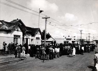 This image shows a large group of people gathered in the High Street of Hastings listening to a speaker on a podium. A double fronted timber building with a sign “Hastings Hall” is in the left foreground and a line of shops stretch into the background. The Australian flag is flying above the hall.