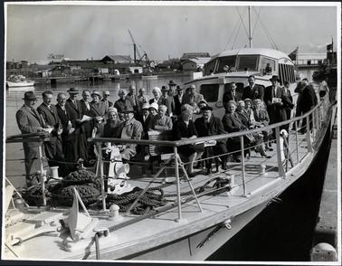 This image shows a large group of people standing and sitting on the deck of a boat beside a jetty.  Twenty-one people are standing and twelve people are sitting.  A crew member is standing at the rail of the boat on the far right-hand side of the photograph.  Everyone is very well dressed and all are wearing coats.  There is a flag flying at the back and large ropes are at the front of the boat.  Small boats, buildings and a crane can be seen in the background.  Two of the buildings are displaying large signs on their roofs - ‘Laconia’ and ‘Johnsons Tyne Foundry’. 