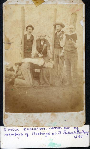 This image shows a group of five men carrying out a mock execution.  Four men are standing and one is kneeling with his head on a block and his hands behind his back.  One man is leaning over him with a club in his hand.  A small dog is on the left-hand side of the image.  The man on the right-hand side is reading from a book and pointing his finger.  Two men are wearing ordinary hats, and two are wearing ‘executioners’ hats.  A piece of cardboard is taped to the bottom and reads: “A mock execution carried out by members of Hastings 40 Pr Bullock Battery 1895”