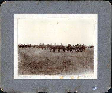 This sepia image is mounted onto grey coloured mount board and depicts the Hastings Battery in camp on 01/04/1907. It shows men in uniform on horseback plus mounted guns on wheels attached to men on horses .  A man dressed in dark pants, jacket and white shirt wearing a hat is standing in front of the gun.  Tents can be seen in the background on right-hand side.  Grass in the foreground. Written on bottom of photo “McAlpine Bros” and “12 Cato St Prahran, Melb April 1. 1907” On the back written in ink “42c Hastings in Battery Camp 1907” “J White Bending over” Written in pencil “1/6 each”.