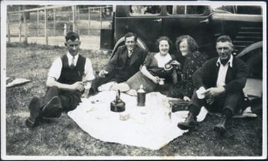 This image shows a group of three men and two women sitting on the grass enjoying a picnic.  A motor vehicle is behind them.  The man on the left is wearing a white shirt with the sleeves rolled up, a tie and vest.  The man beside him is wearing a three piece suit and the man on the right-hand side is wearing a suit with no tie.  The woman on the left has a wide white collar on her dress and the older woman is wearing a print dress.  A white table cloth is in front of the group set for a picnic, including a kettle.  A person can be seen on a tennis court behind a high wire fence in the background and behind the court someone is sitting on a garden chair.  Another garden chair can be seen to his right.  